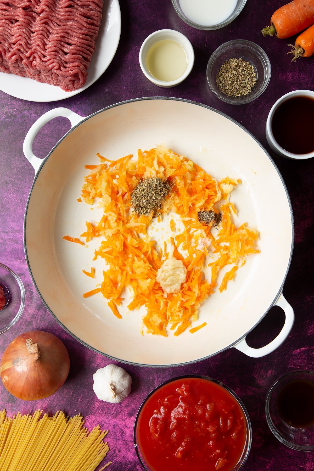 A large frying pan with fried carrot and onion, plus minced garlic and dried oregano. Ingredients to make spaghetti bolognese Gordon Ramsay style surround the pan.