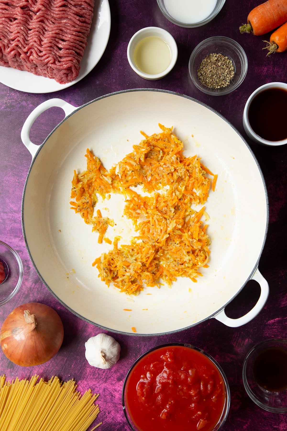 A large frying pan with fried carrot, onion, garlic and dried oregano. Ingredients to make spaghetti bolognese Gordon Ramsay style surround the pan.