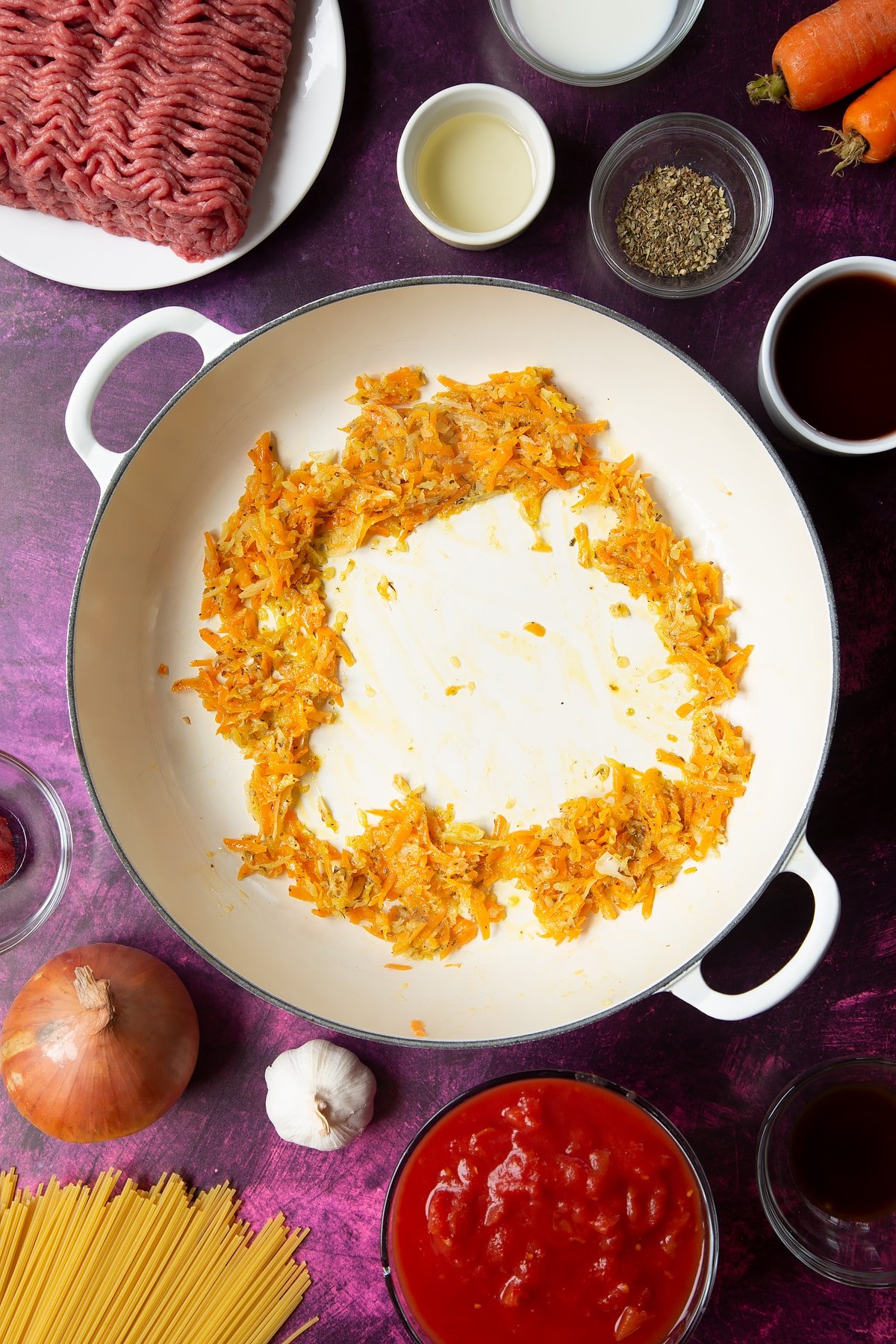 A large frying pan with fried carrot, onion, garlic and dried oregano pushed to the sides of the pan. Ingredients to make spaghetti bolognese Gordon Ramsay style surround the pan.