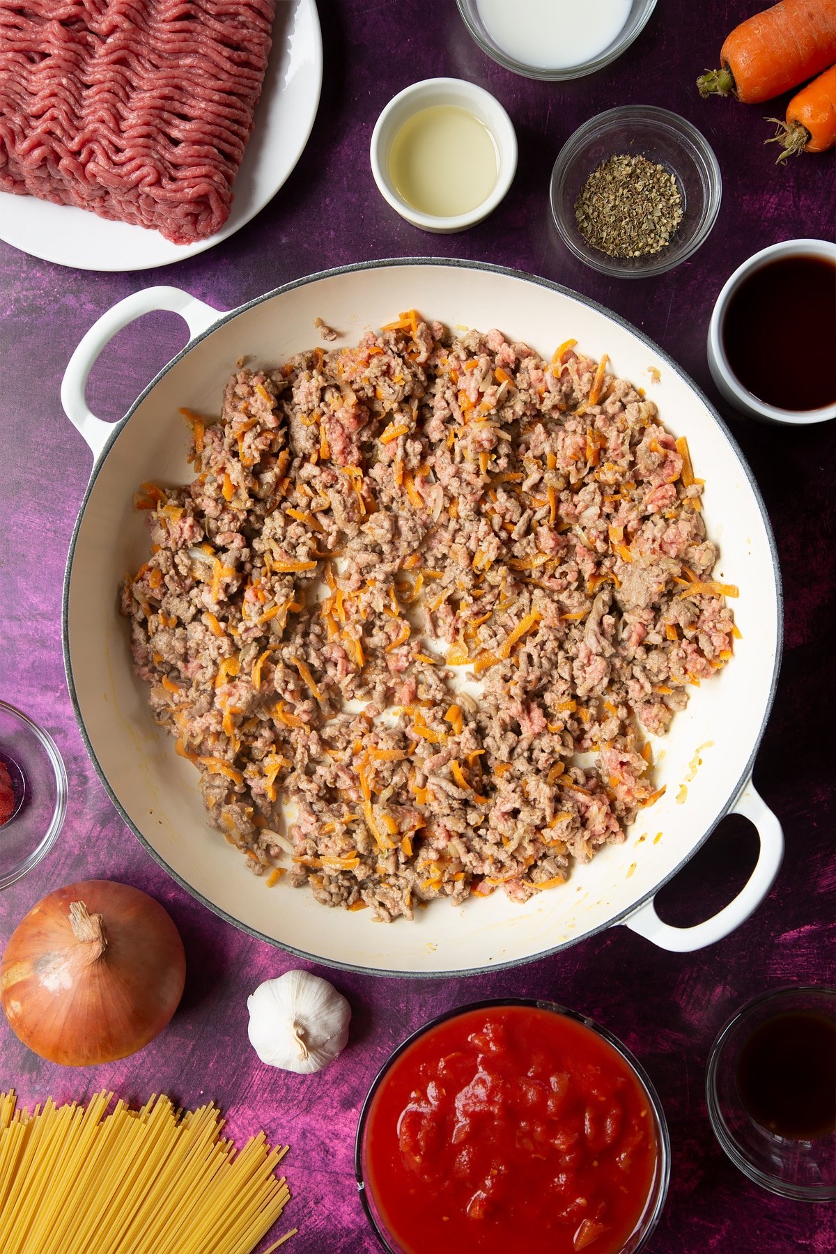 A large frying pan with fried carrot, onion, garlic, dried oregano and lean beef mince. Ingredients to make spaghetti bolognese Gordon Ramsay style surround the pan.