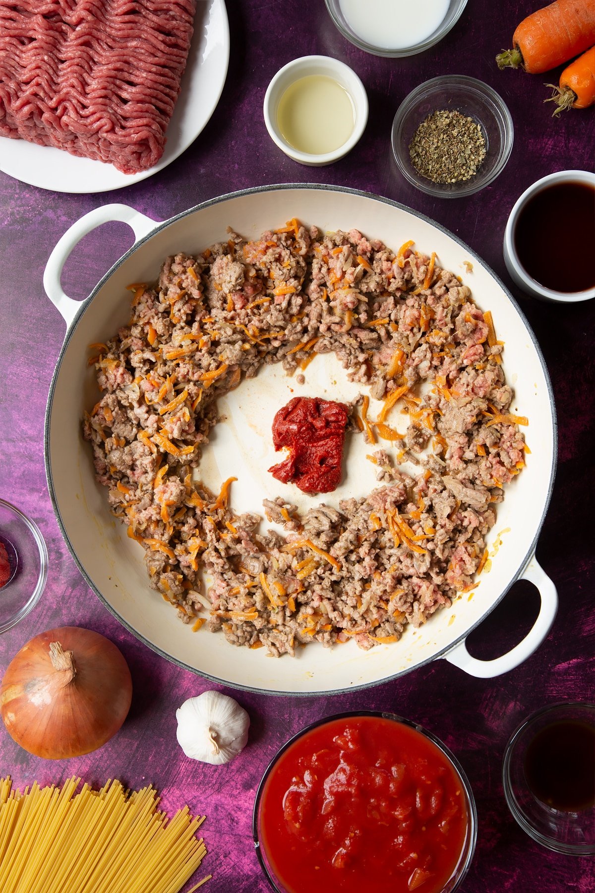 A large frying pan with fried carrot, onion, garlic, dried oregano and lean beef mince. Tomato puree has been added to the centre of the pan. Ingredients to make spaghetti bolognese Gordon Ramsay style surround the pan.