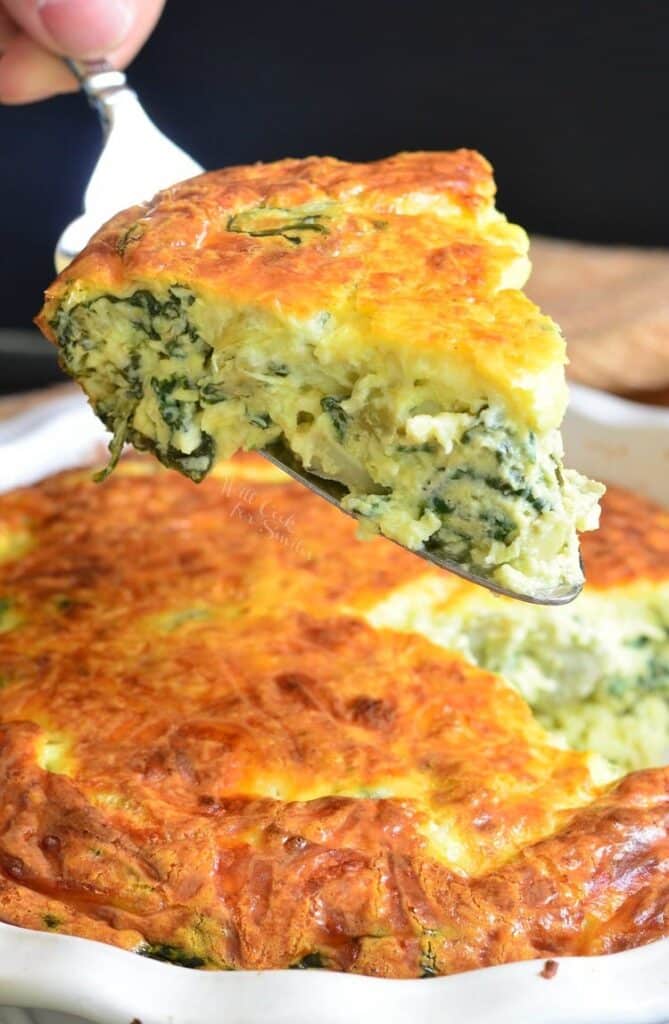 A slice of Spinach Artichoke Crustless Quiche being picked up using a serving fork. The remainder of the quiche is seen below the slice inside a dish.