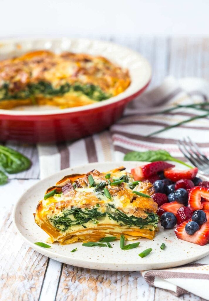 A slice of bacon and cheddar quiche on a decorative plate served with strawberries and blueberries. The large dish of bacon and cheddar quiche sits in the background.