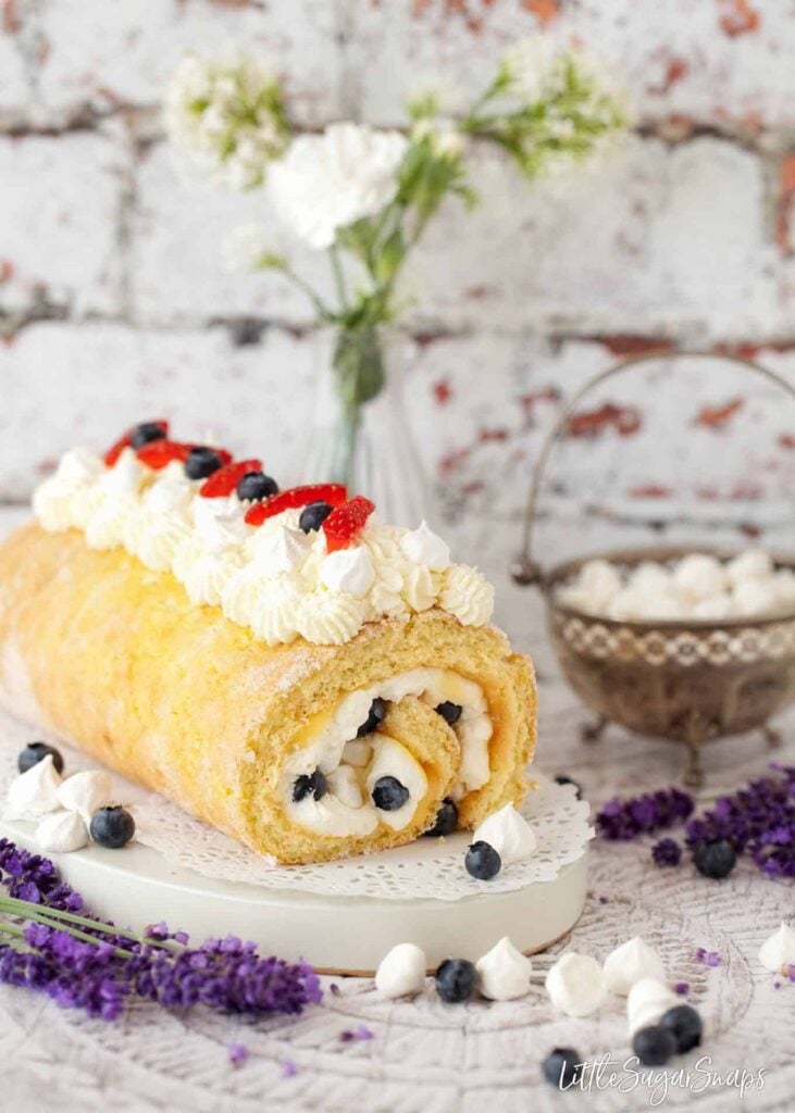 A lemon meringue Swiss roll with summer berries sit on a decorative plate on top of a white table. The area is decorated with miniature meringues, blueberries and lavender.