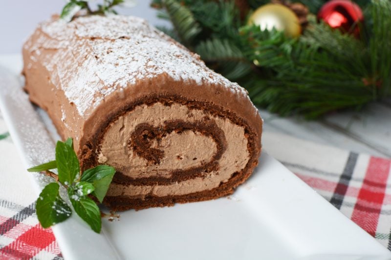 Large yule log cake sitting on a long rectangular plate with a red, white and black checked tea towel underneeth. In the background there's some foliage with christmas baubles. 