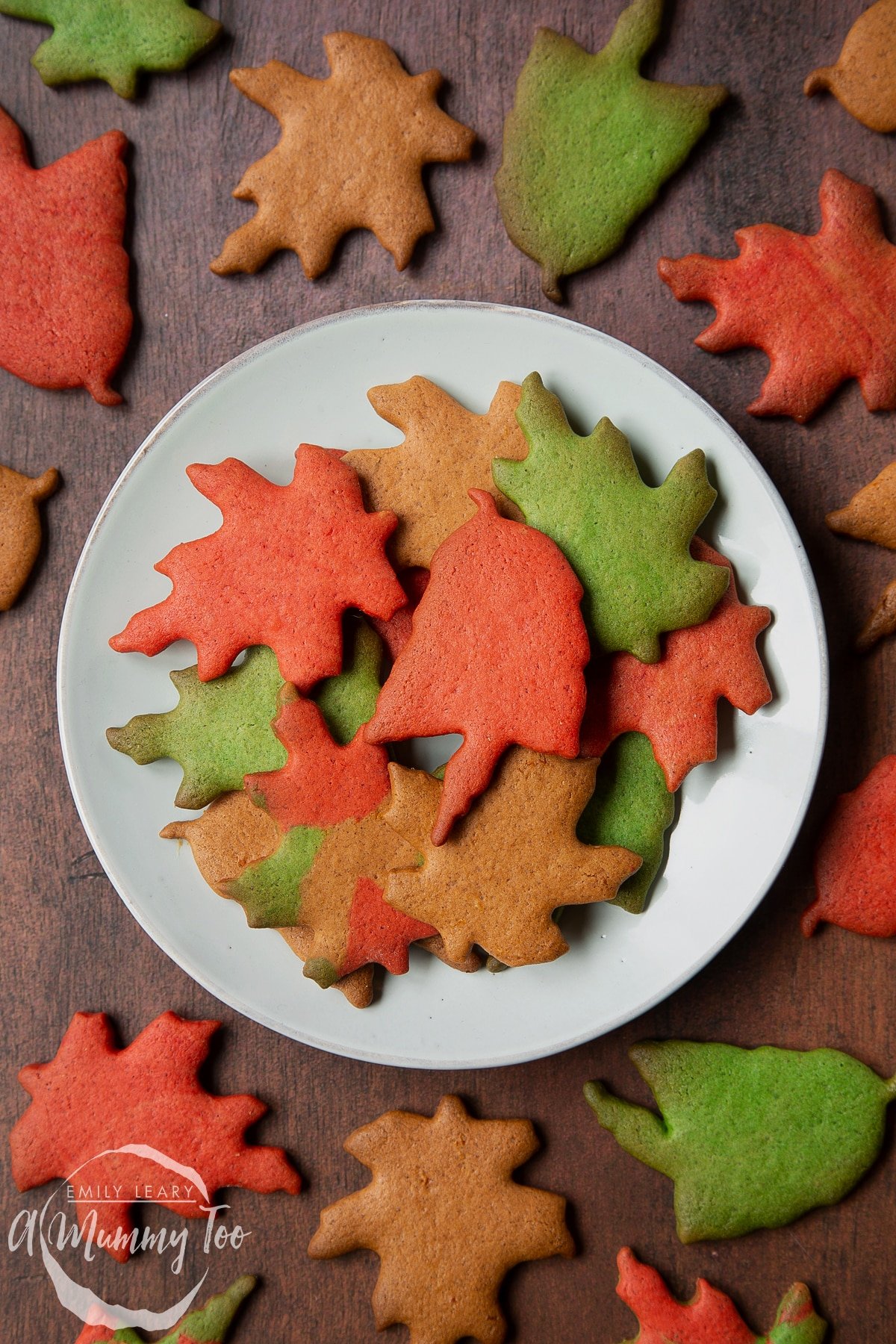 A plate of colourful red, green and brown autumn cookies cut into the shapes of autumn leaves. A red leaf biscuit sits in the centre.