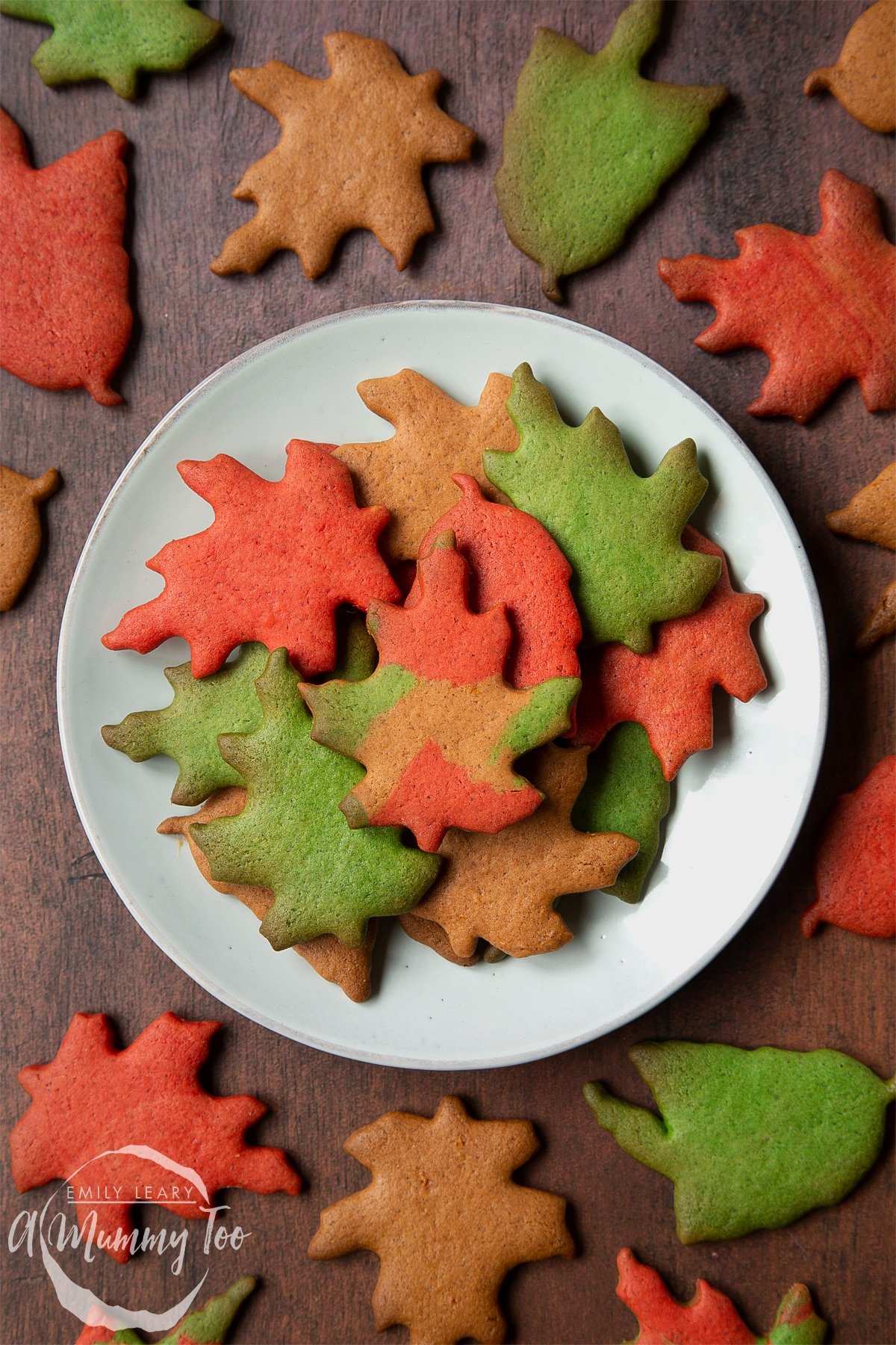 A plate of colourful red, green and brown autumn cookies cut into the shapes of autumn leaves. More Autumn biscuits are arranged on the wooden surface surrounding the plate.