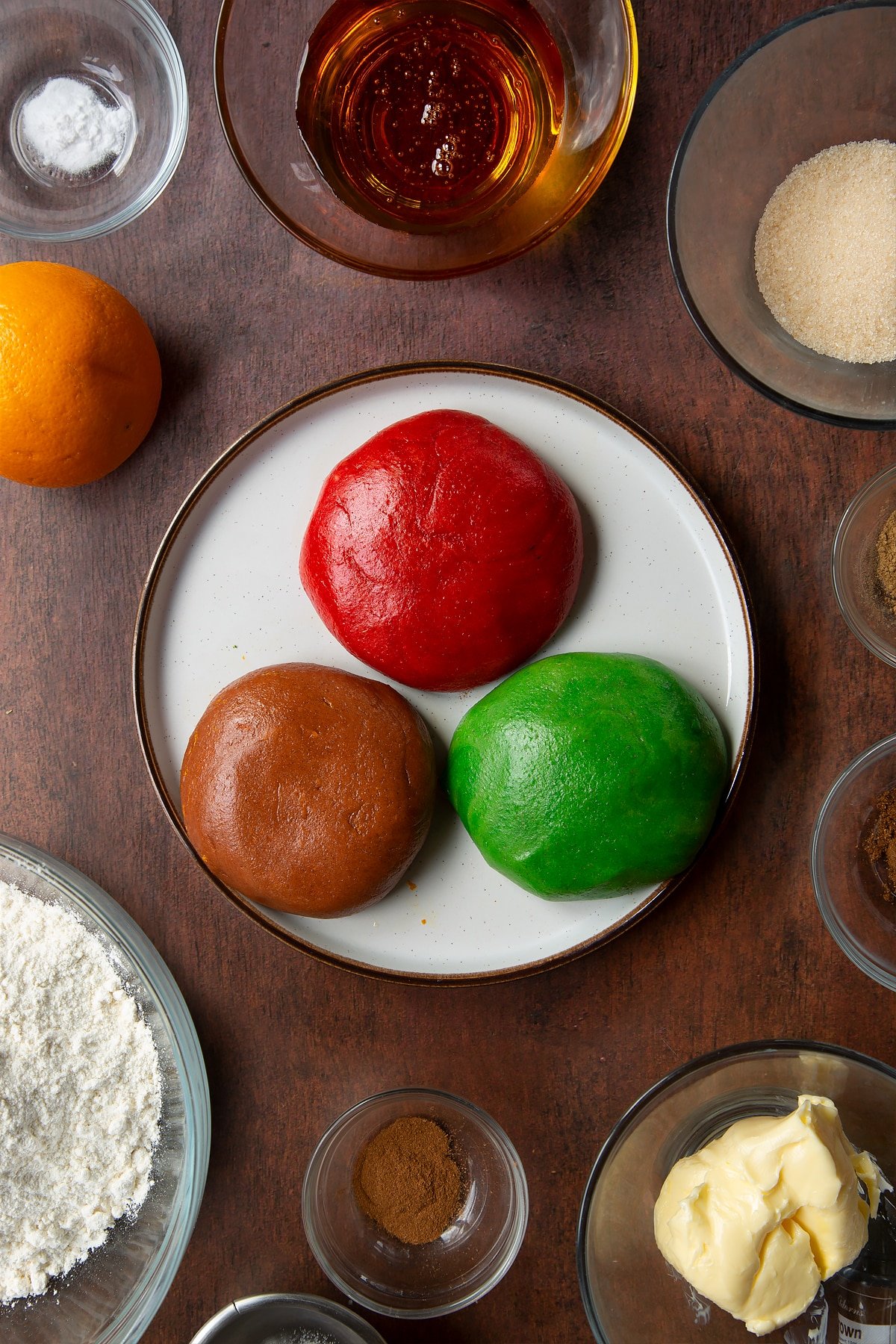 Three balls of coloured and spiced cookie dough on a plate. Ingredients to make autumn cookies surround the plate.