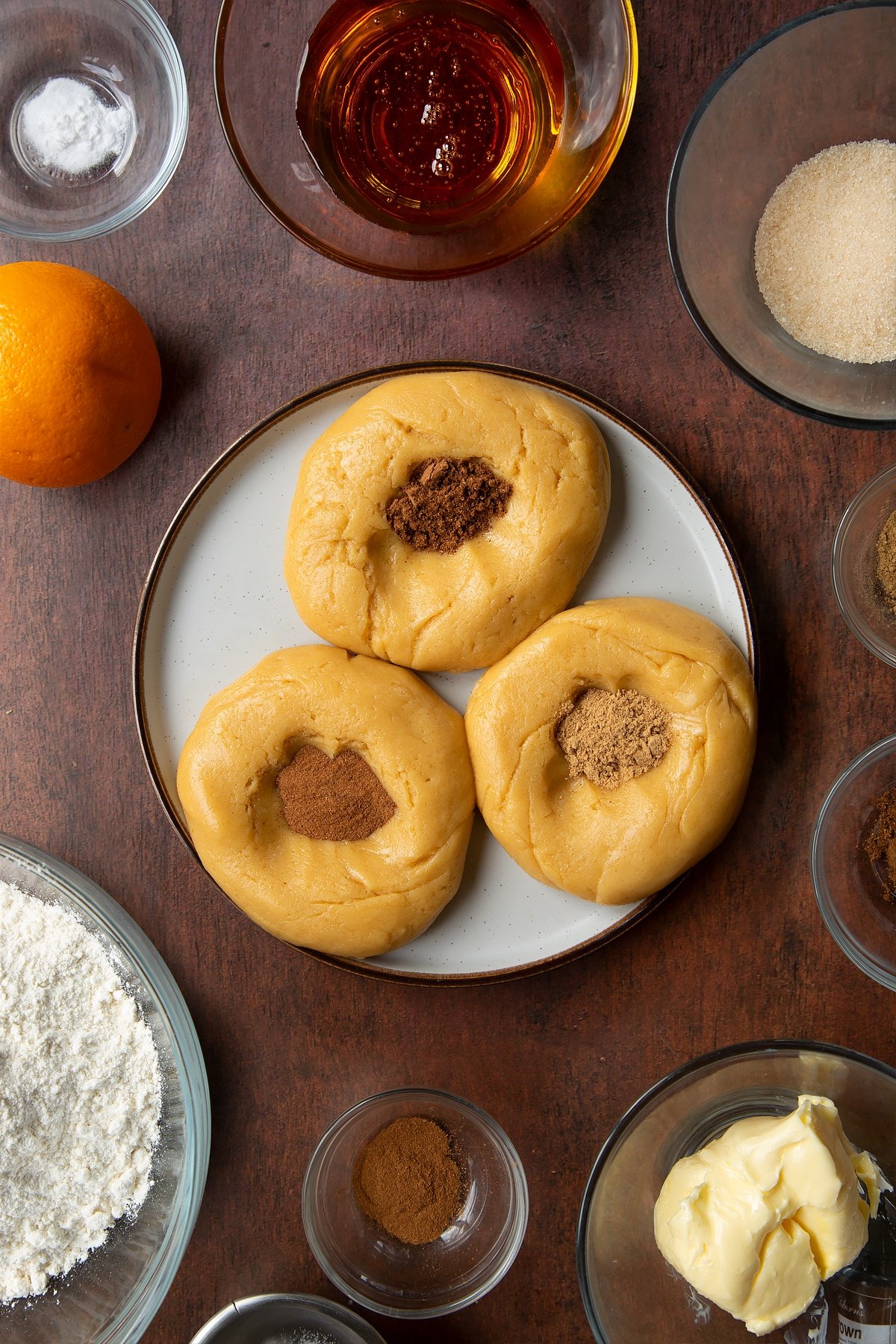 Three balls of cookie dough on a plate with spices on top of them. Ingredients to make autumn cookies surround the plate.