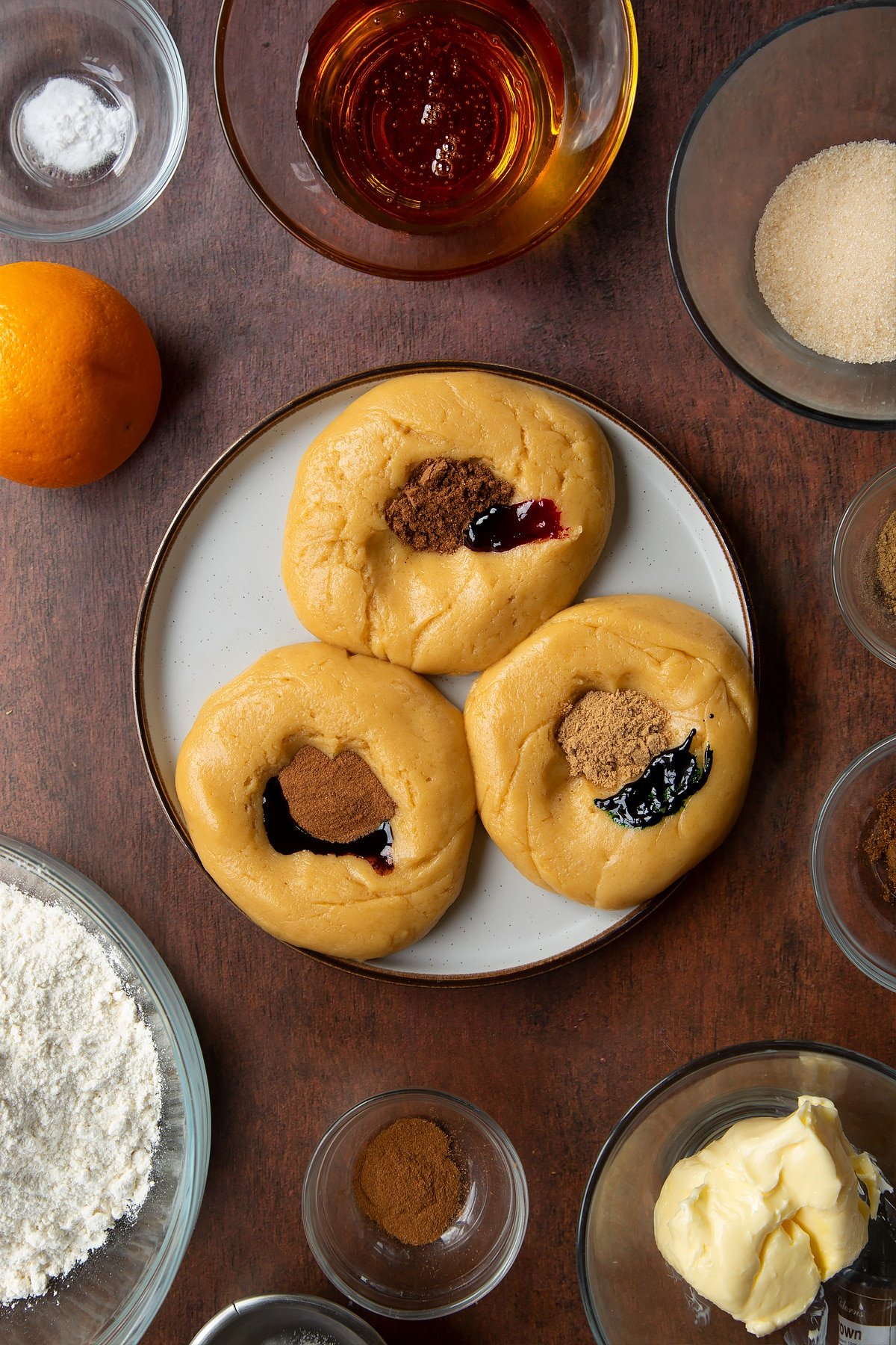 Three balls of cookie dough on a plate with spices and food coloured on top of them. Ingredients to make autumn cookies surround the plate.