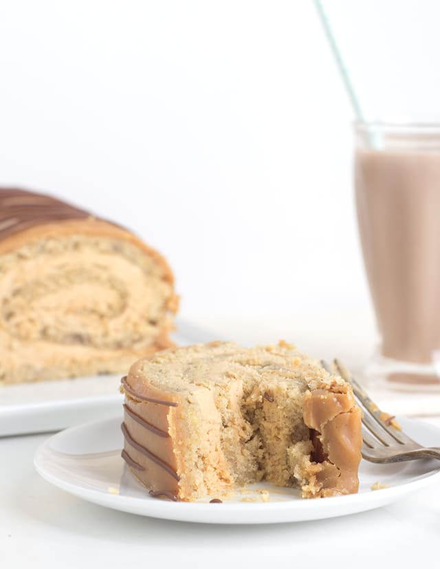 A slice of Banana Roll Cake With Whipped Peanut Butter Ganache sits on a white plate with a fork on the side. In the background there's a milkshake.
