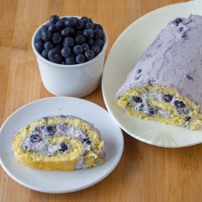 Blueberry Swiss roll cake with the main cake sitting on a large decorative plate and a slice placed on white circular plate with a cup of blueberries at the side. All sitting ontop of a wooden surface.