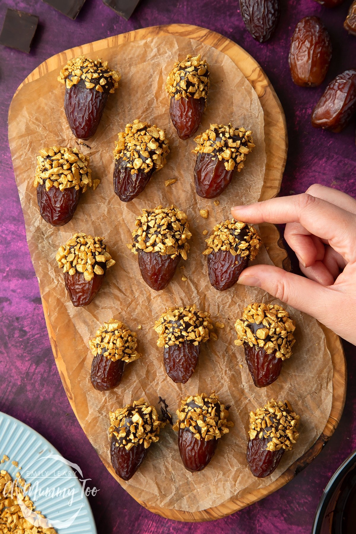 Medjool dates dipped in chocolate. The chocolate dates are on a wooden board lined with brown baking paper. The dates have be studded with gold chopped nuts. A hand reaches to take one.