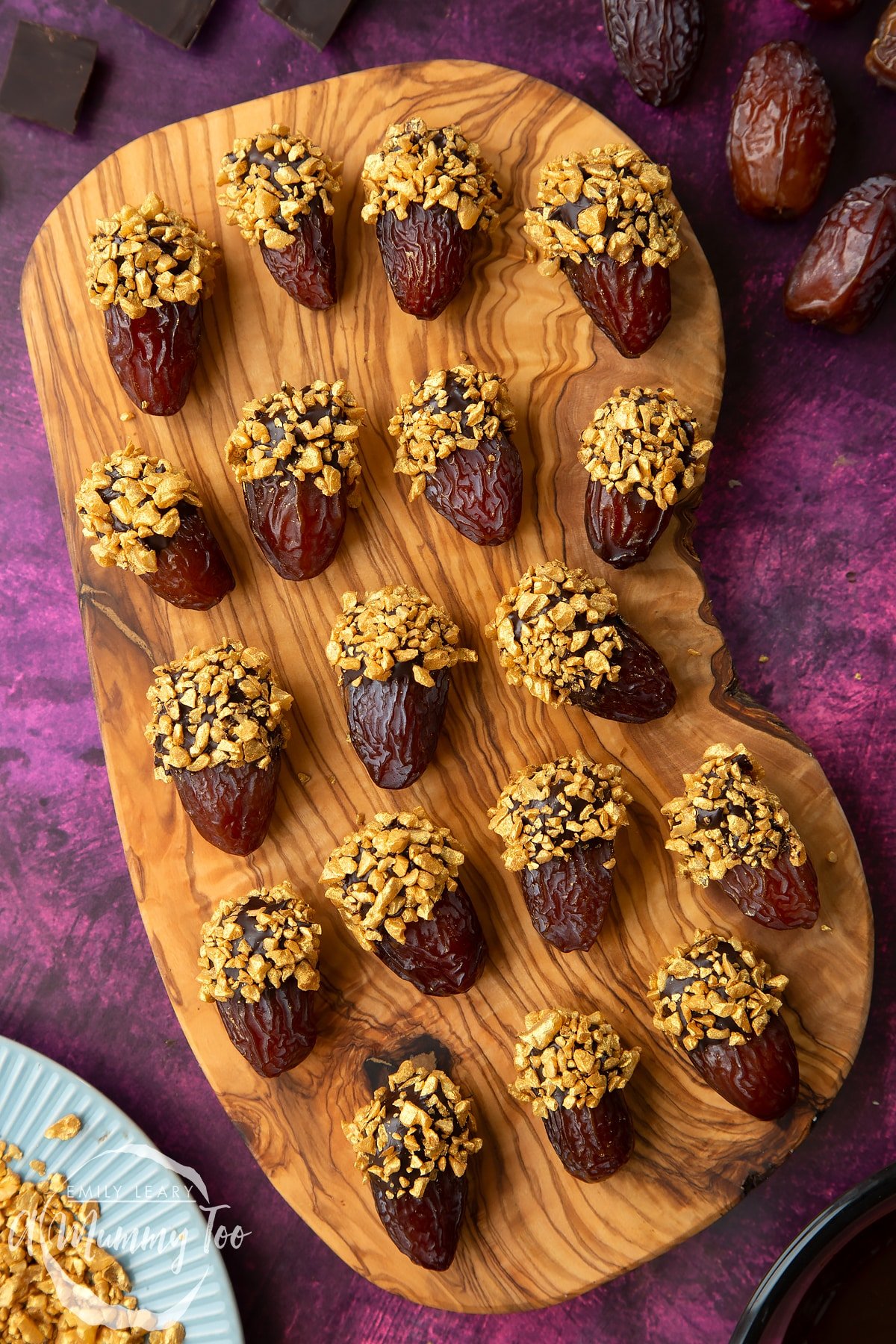 Medjool dates dipped in chocolate. The chocolate dates are on a wooden board and have be studded with gold chopped nuts. Nuts and chocolate surround the board.