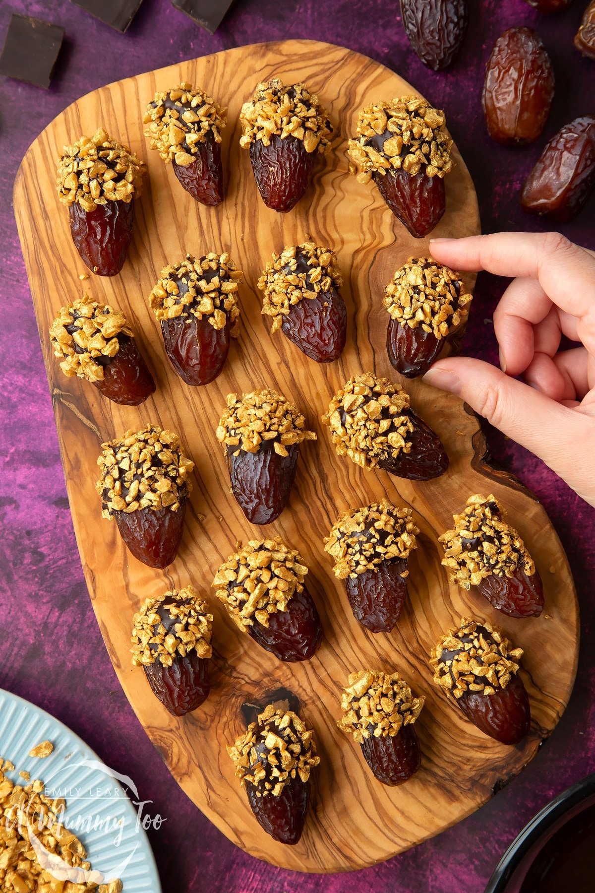 Medjool dates dipped in chocolate. The chocolate dates are on a wooden board and have be studded with gold chopped nuts. Nuts and chocolate surround the board. A hand reaches to take a date.