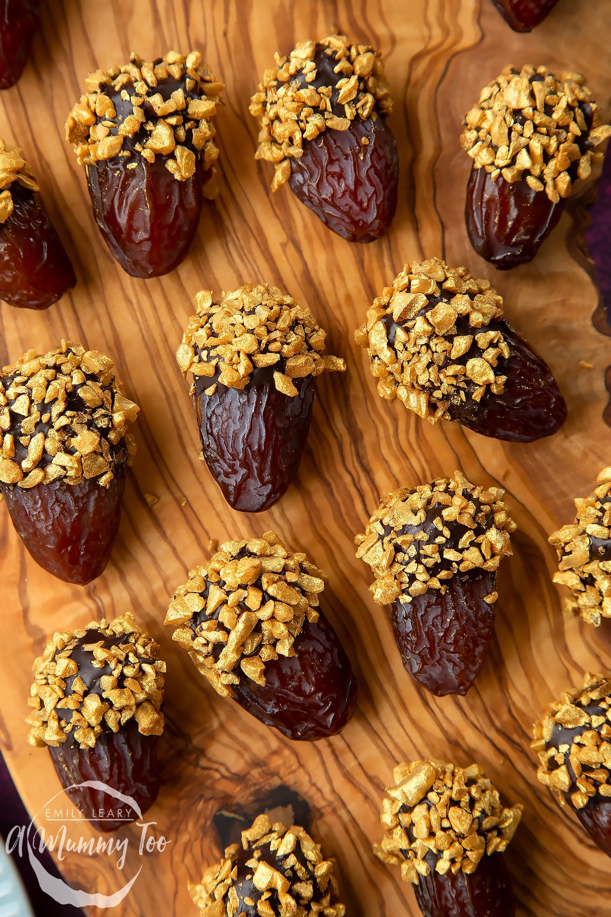 Close up of medjool dates dipped in chocolate. The chocolate dates are on a wooden board and have be studded with gold chopped nuts.