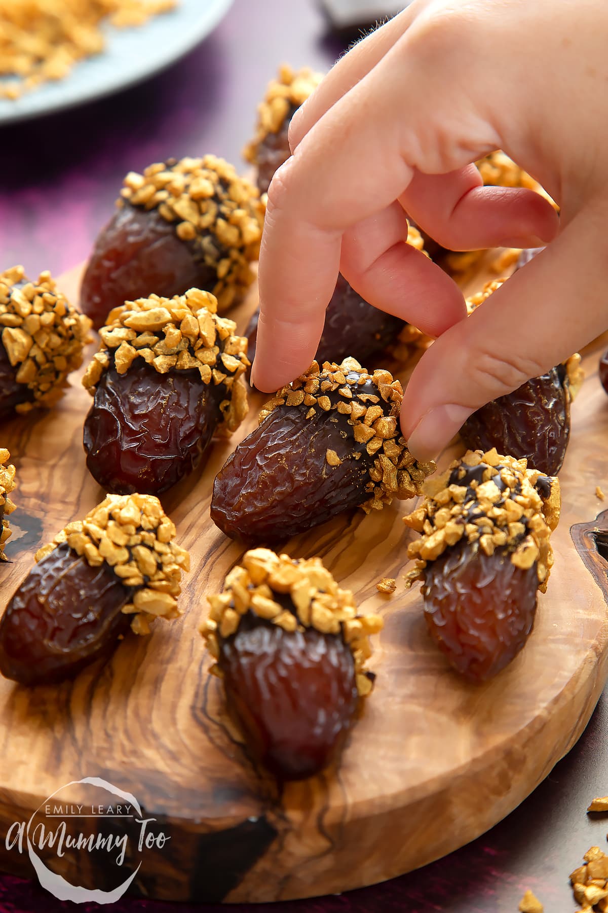 Medjool dates dipped in chocolate. The chocolate dates are on a wooden board and have be studded with gold chopped nuts. A hand reaches to take a date.