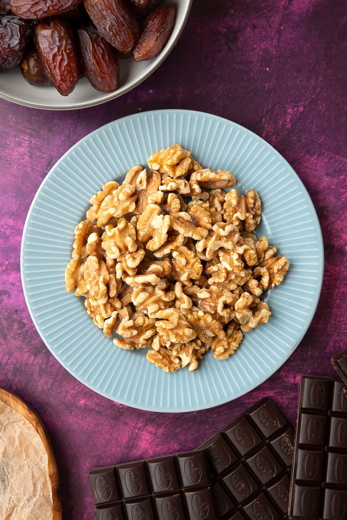 Walnut halves on a blue plate, surrounded by ingredients to make chocolate dates. 