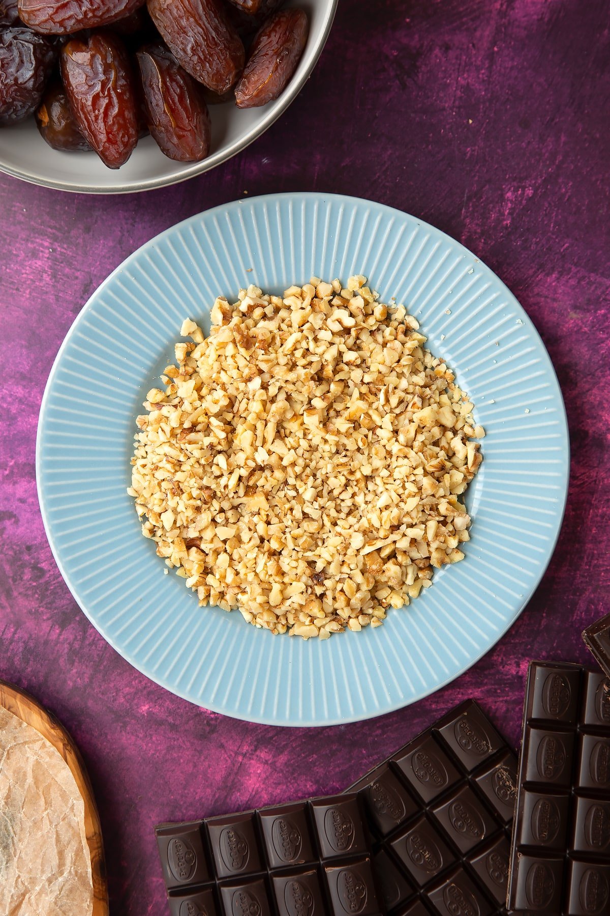 Chopped walnuts on a blue plate, surrounded by ingredients to make chocolate dates. 