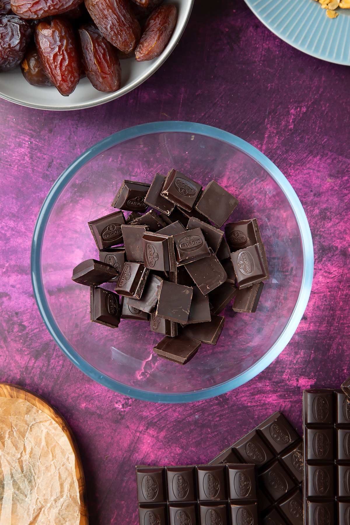 Dark chocolate squares in a bowl, surrounded by ingredients to make chocolate dates. 