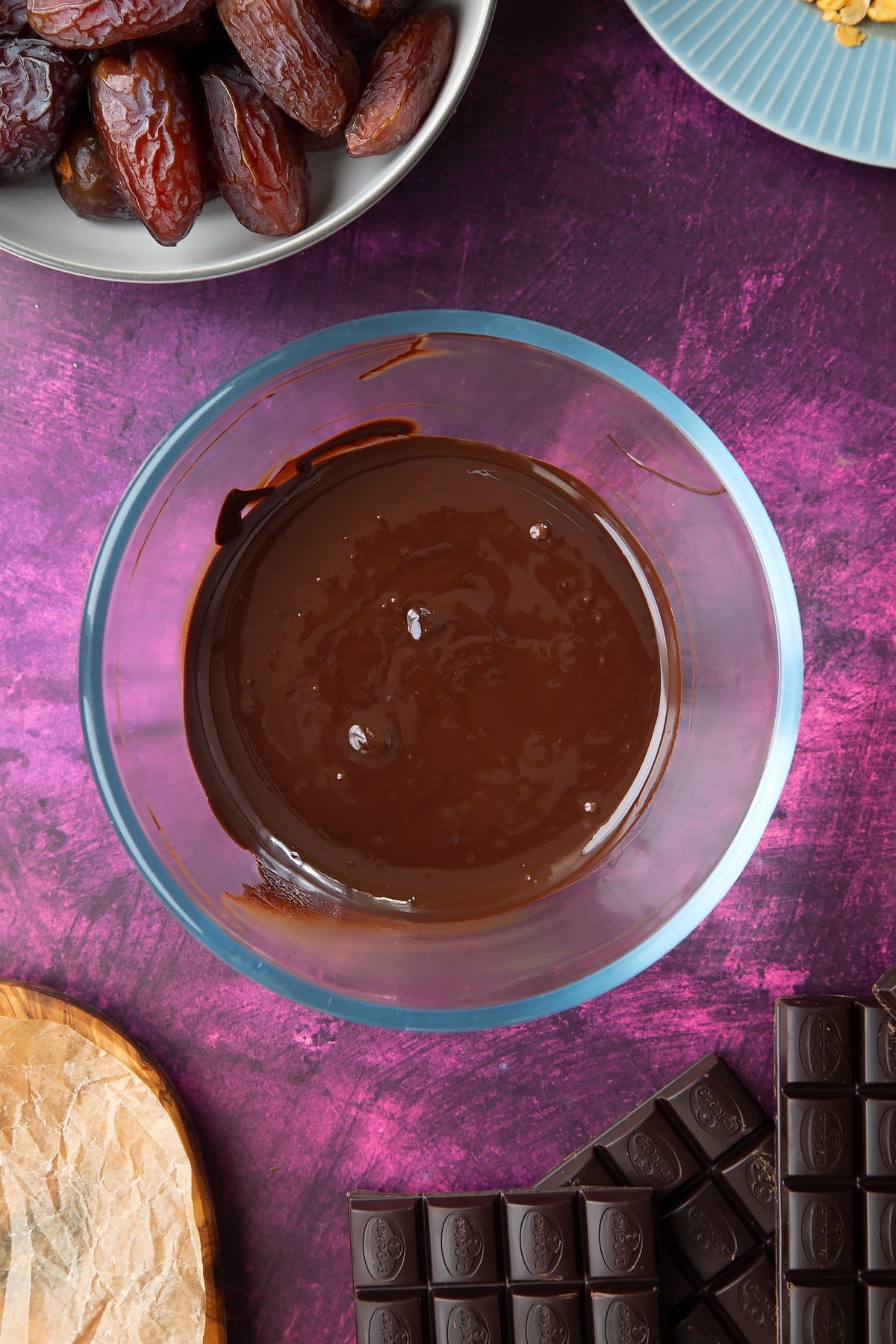 Melted chocolate in a bowl, surrounded by ingredients to make chocolate dates. 