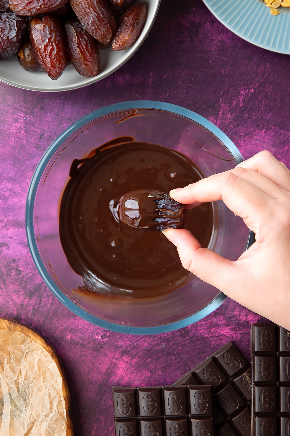 Melted chocolate in a bowl, surrounded by ingredients to make chocolate dates.  A hand holds a date above the bowl, which is half coated with chocolate.