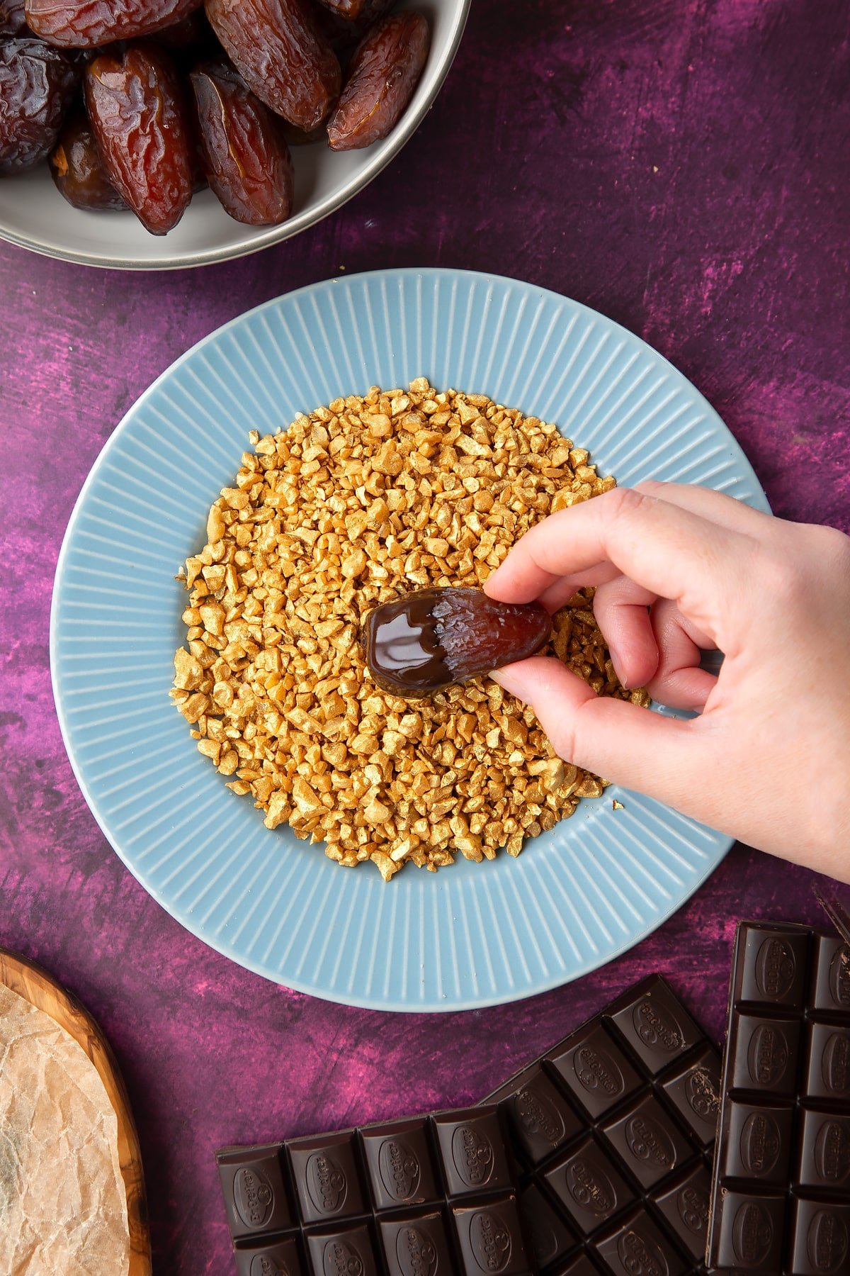 Chopped walnuts sprayed gold on a blue plate, surrounded by ingredients to make chocolate dates. A hand dips a chocolate coated date into the nuts.