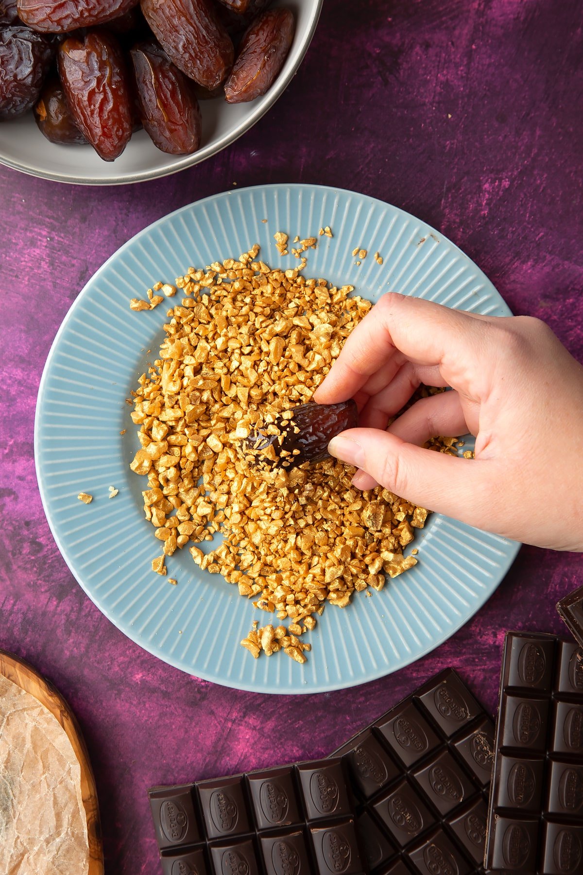 Chopped walnuts sprayed gold on a blue plate, surrounded by ingredients to make chocolate dates. A hand holds a chocolate and nut coated date.