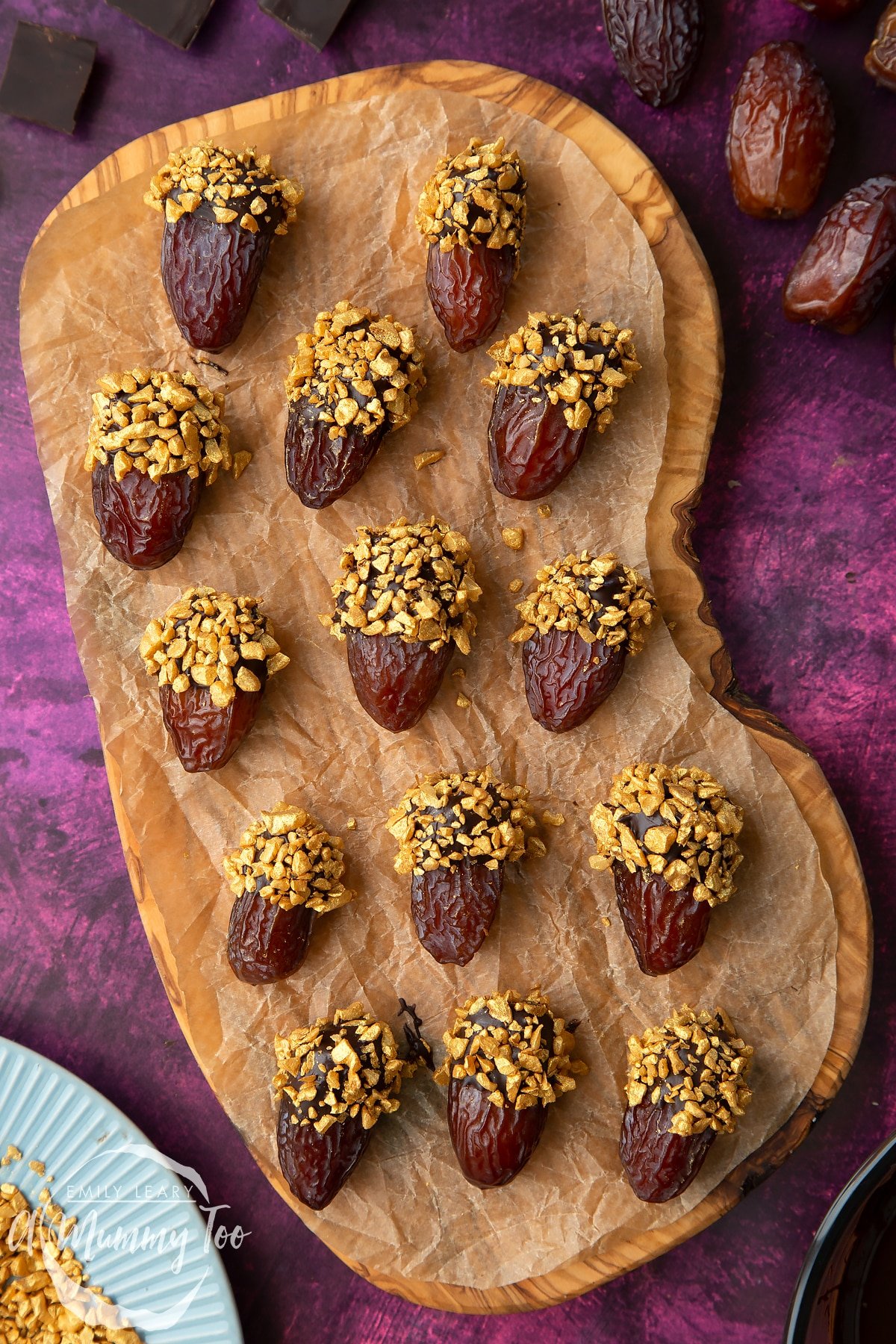 Medjool dates dipped in chocolate. The chocolate dates are on a wooden board lined with brown baking paper. The dates have be studded with gold chopped nuts. Nuts and chocolate surround the board.