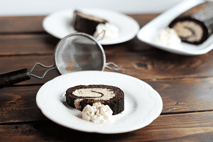 A wooden table has two plates with a slice of chocolate rolled cake with cookie dough. In the background you can see a rectangular plate with the remainder of the chocolate rolled cake. 