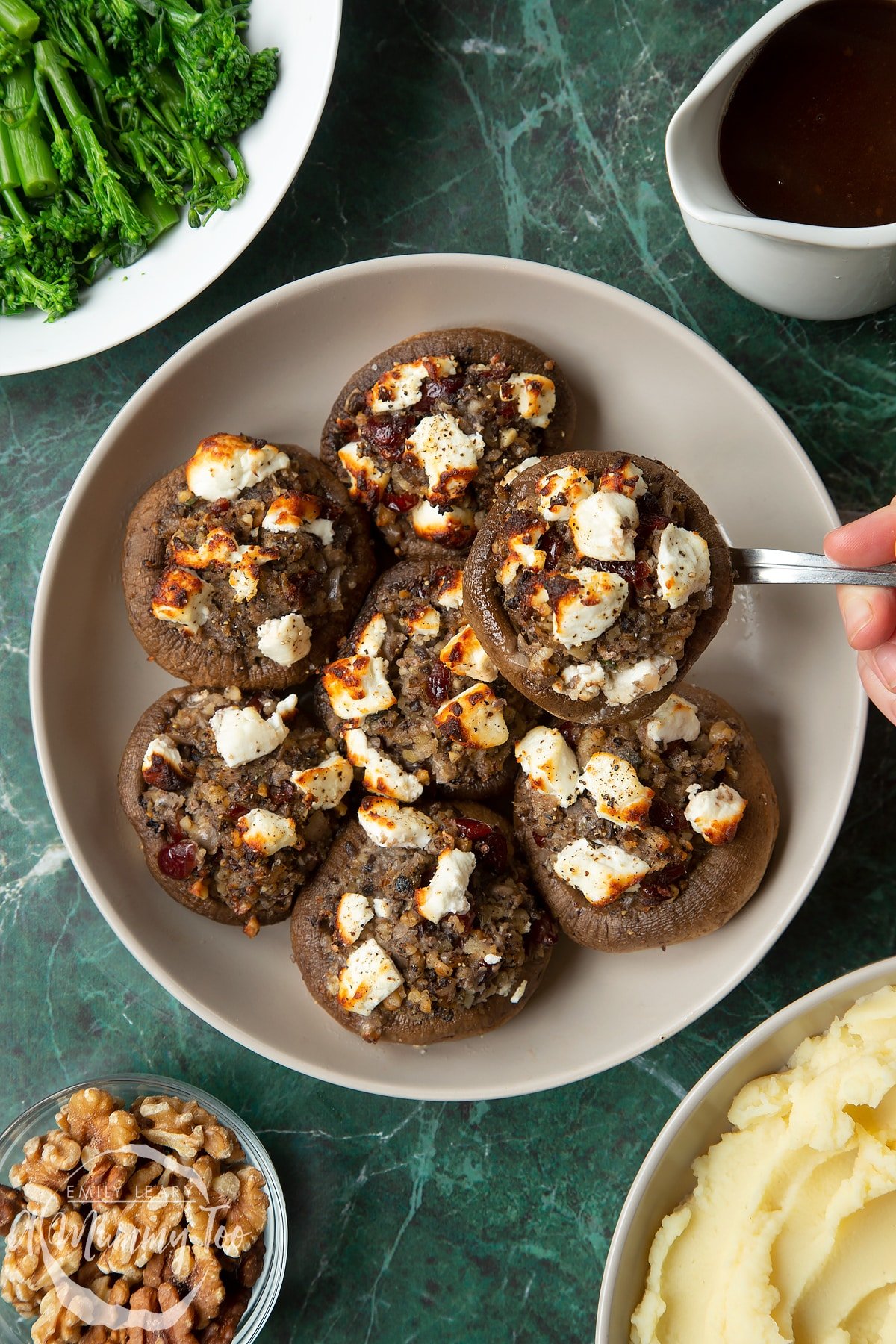 Overhead shot of a hand picking up a festive stuffed mushrooms recipe served on a gray plate with a mummy too logo in the lower-right corner