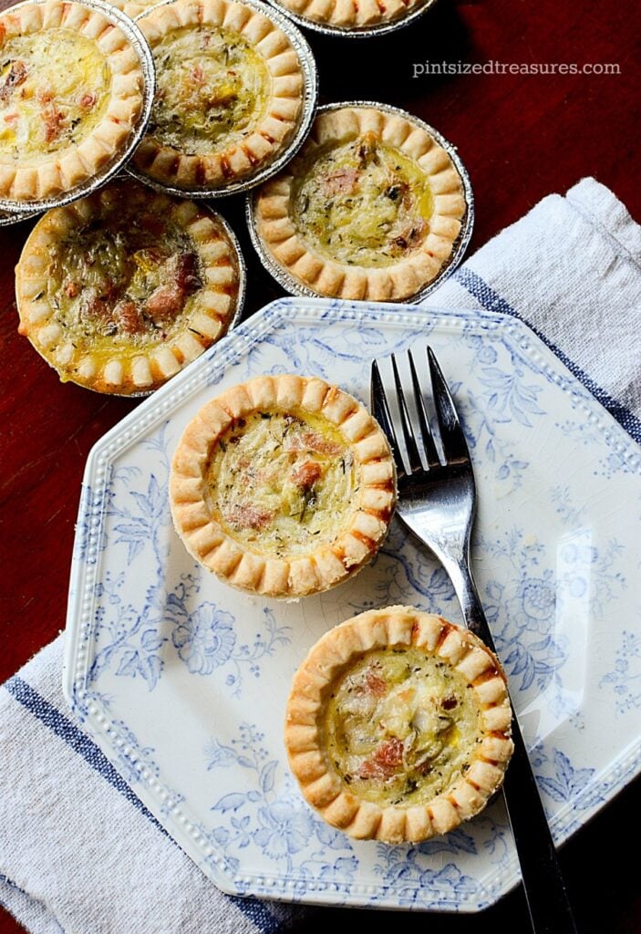 Two Ham & Havarti Mini Quiches sit on a decorative blue and white plate with a fork at the side. In the top right of the image there's a stack of additional miniture Ham & Havarti Mini Quiches.