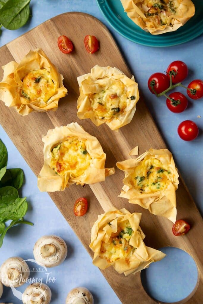 Four Filo Pastry Mini Quiches laid on a wooden chopping board on a light blue background. At the side there's a vine of cherry tomatoes and a blue plate with another mini quiche.