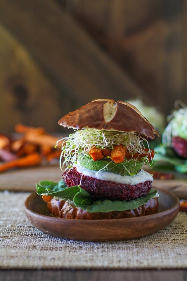 Beetroot burger with multiple veg toppings in a brioche bun on a small wooden plate with vegetables and potato fries in the background.