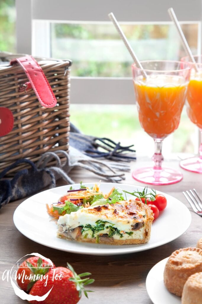 A slice of Chestnut Mushroom, Spinach & Feta Quiche sits on a white plate with a side salad and some on the vine tomatoes. At the front of the image you can see some pork pies on a plate and some strawberries and in the background a picnic basket and two drinks.