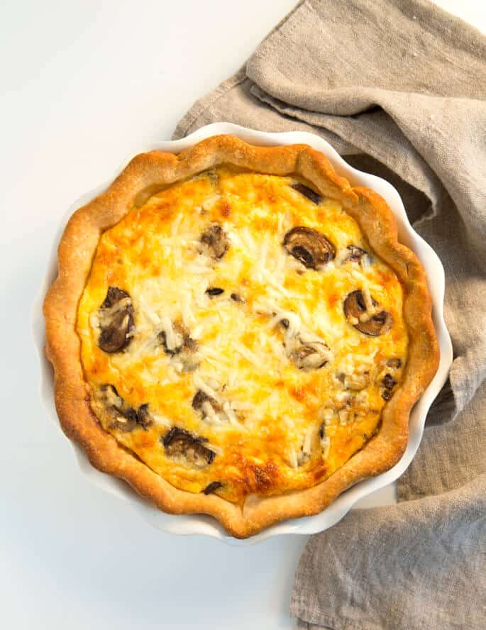 This gluten free quiche sits inside a decorative pie dish on a white background. At the side there's a natural tea towel / cloth. 