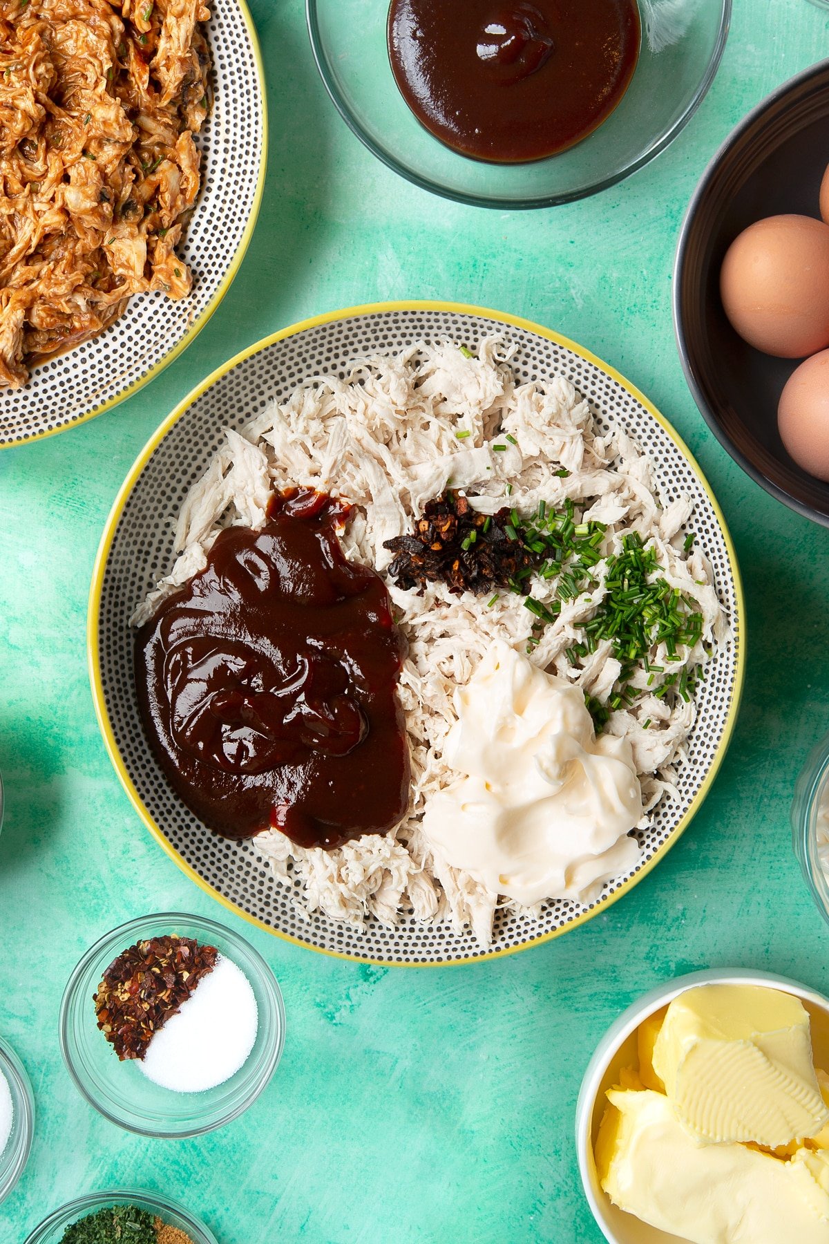 Shredded chicken breast in a bowl, topped with barbecue sauce, mayo, chilli flakes and chopped chives. Ingredients to make chicken doughnuts surround the bowl.