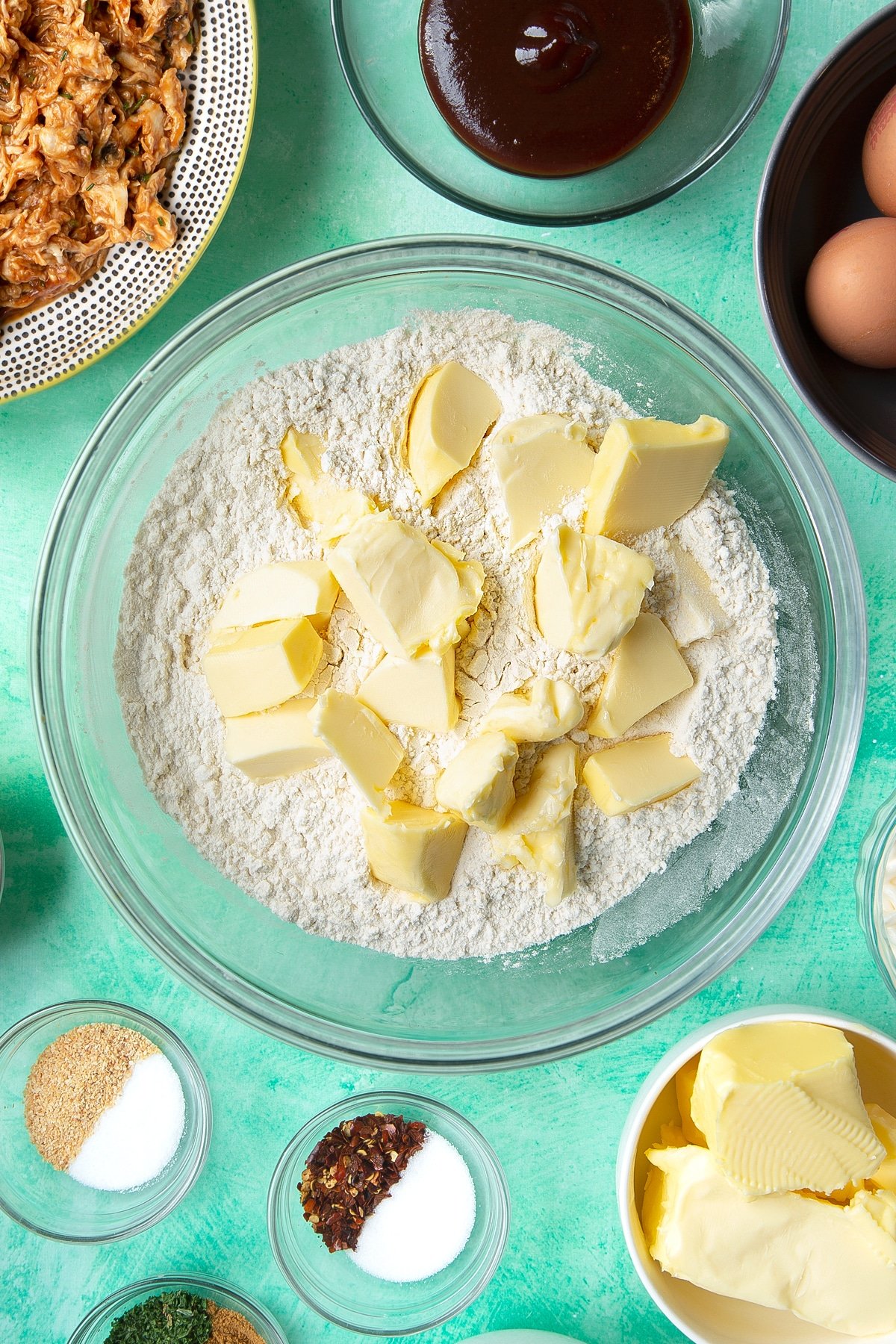 Flour and butter in a glass mixing bowl. Ingredients to make chicken doughnuts surround the bowl.