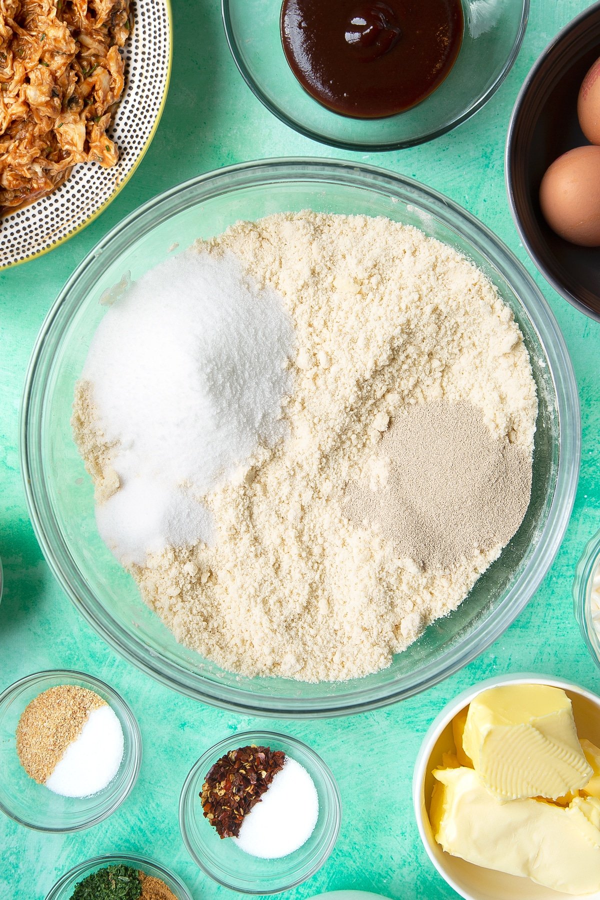 Flour and butter rubbed together in a glass mixing bowl and topped with salt, sugar and yeast. Ingredients to make chicken doughnuts surround the bowl.