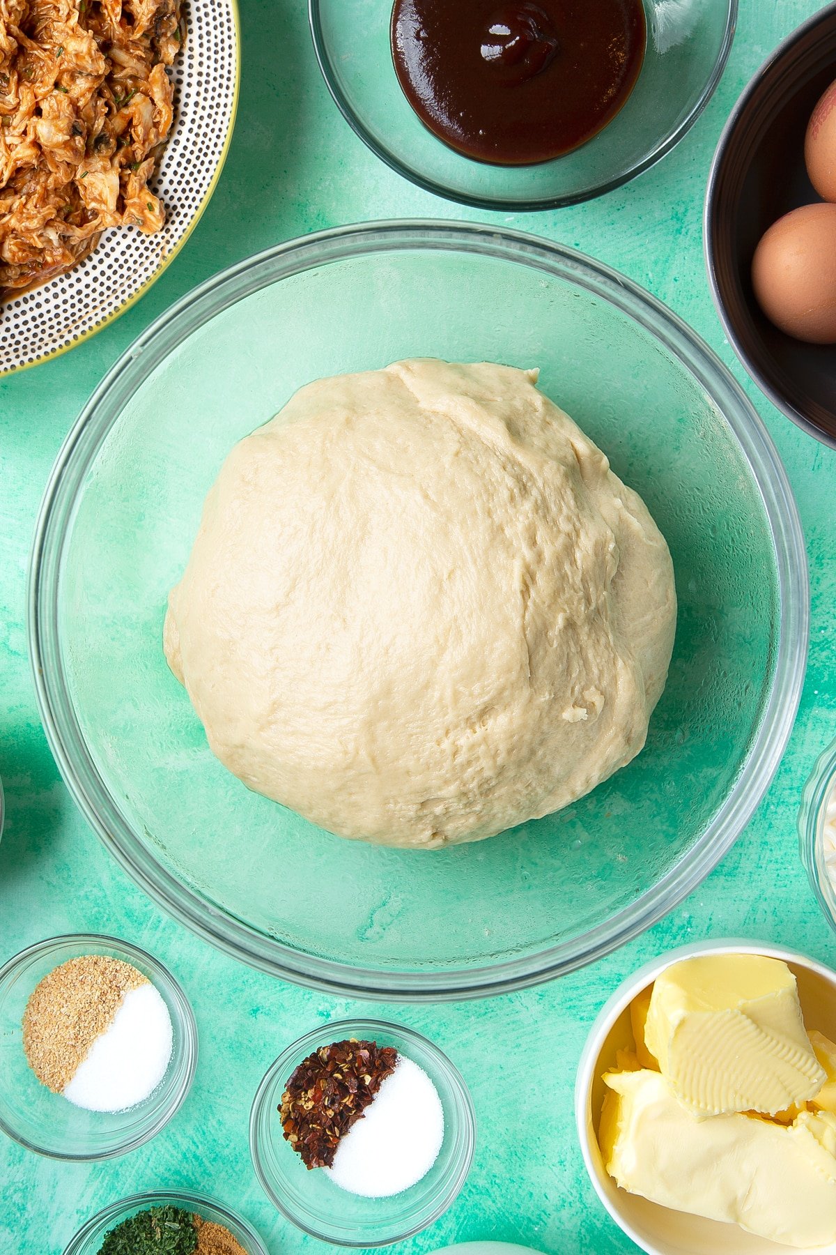 A kneaded and smooth bread dough ball in a glass mixing bowl. Ingredients to make chicken doughnuts surround the bowl.