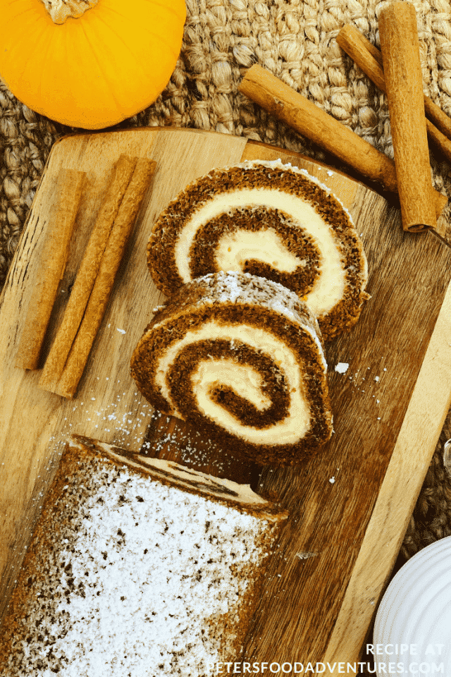 A pumpkin roll with two slices cut off. Cinemon sticks decorate the side of the wooden chopping board next to the roll.