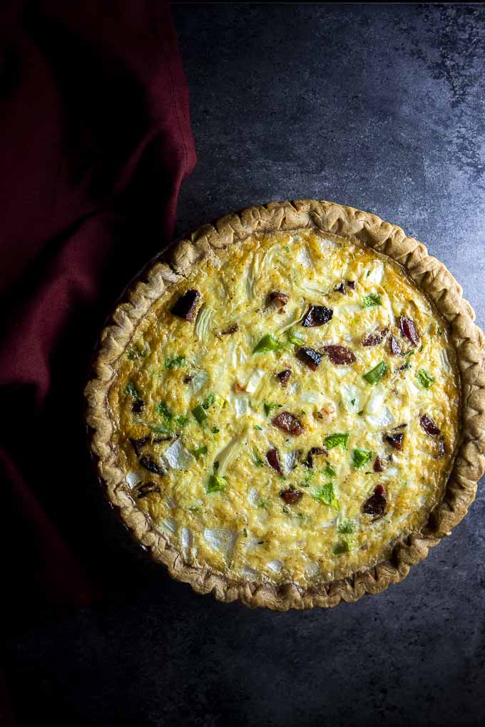 A whole cheesy sausage quiche with shrimp sits on a dark black background with a red napkin at the side.