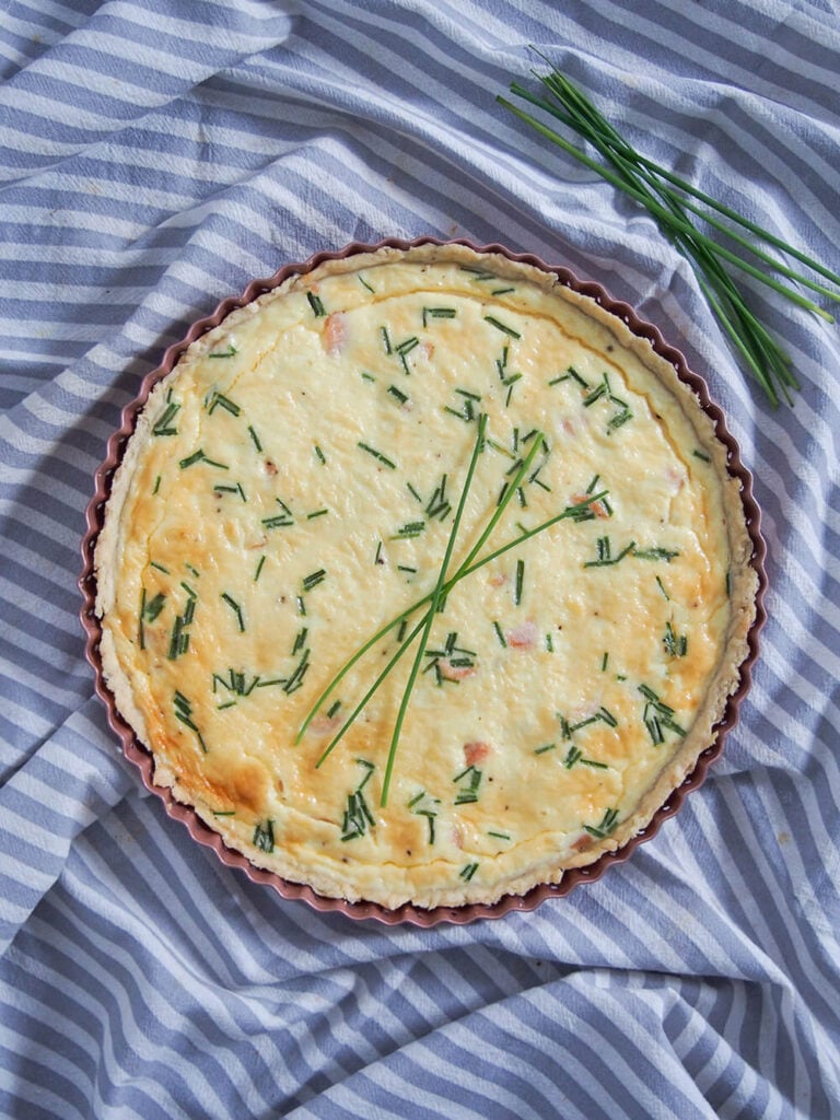 A whole Smoked Salmon Quiche sits inside a tray on a purple and white striped cloth. At the side of the quiche is some unchopped chives.