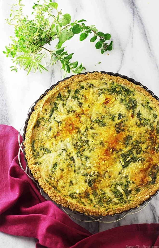 A large Spinach-Gouda Quiche With Quinoa Crust sits on a white marble background, at the side there's some leaves and a red napkin.