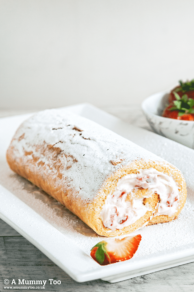A Strawberry and Rosewater Cream Swiss Roll sits on a rectangular plate with a decorative strawberry in the foreground.