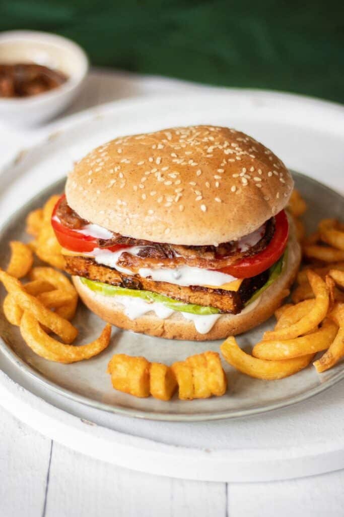 Vegan BBQ Tofu Burger sat on a white / cream plate and surrounded by curly fries. 