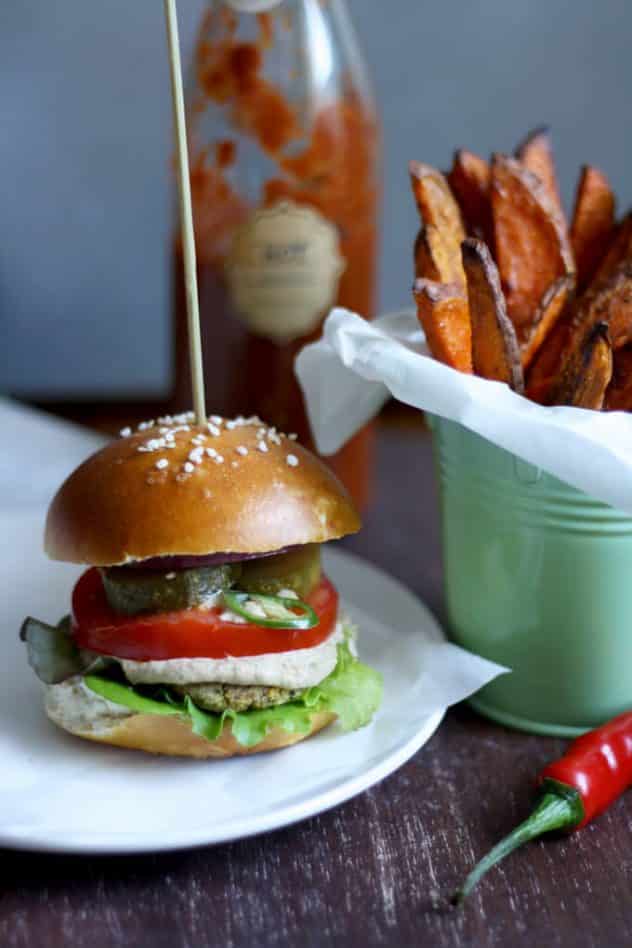 Falafel burger with a skewer going through the top. A bottle of tomato ketchup in the background and a side of fries in the corner. 