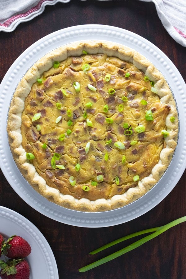 A large vegan quiche lorraine sits on a white plate on a dark wooden table. At the top of the image is a napkin, and at the bottom a plate of strawberries.