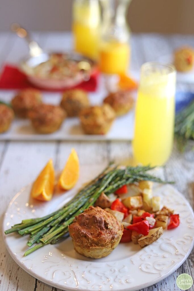 A mini vegan quiche cooked in the airfryer sits on a highly decorative plate with a side of asparagus with chopped potatoes and peppers. Additional mini quiches are on a serving plate in the background.