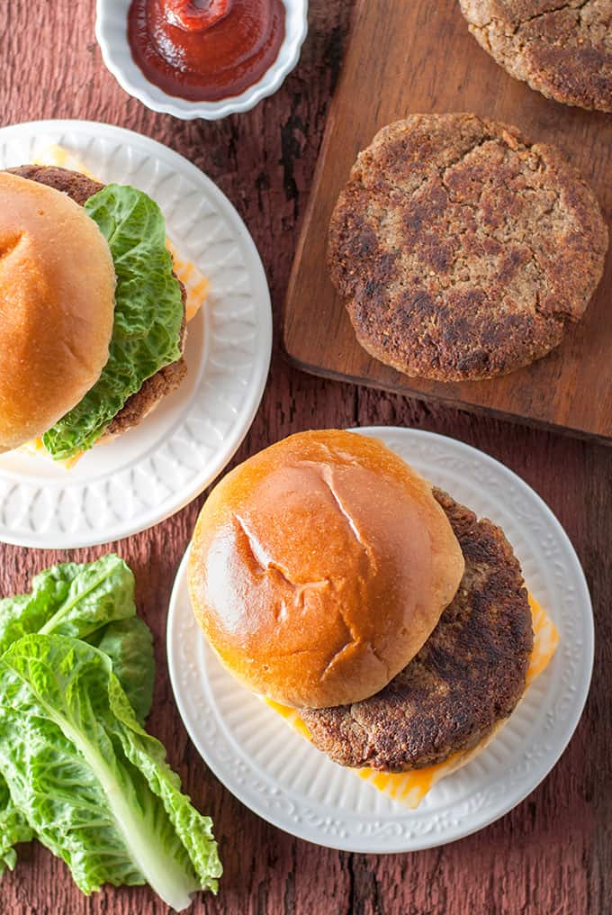 Two walnut burgers on white plates topped with a glazed bun, meanwhile another single burger sits on a wooden chopping board.